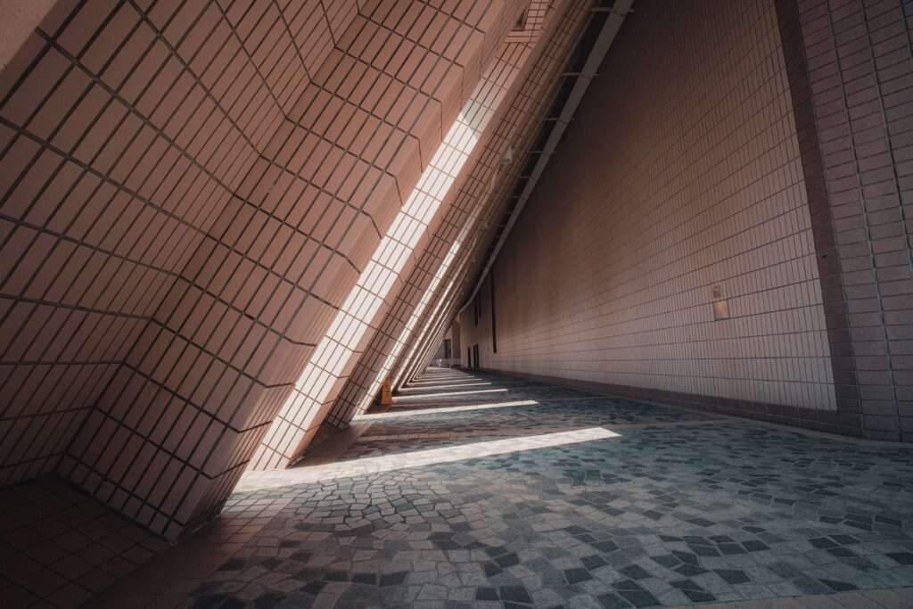 A modern corridor with slanted walls and intricate tile flooring, illuminated by sunlight casting geometric patterns.