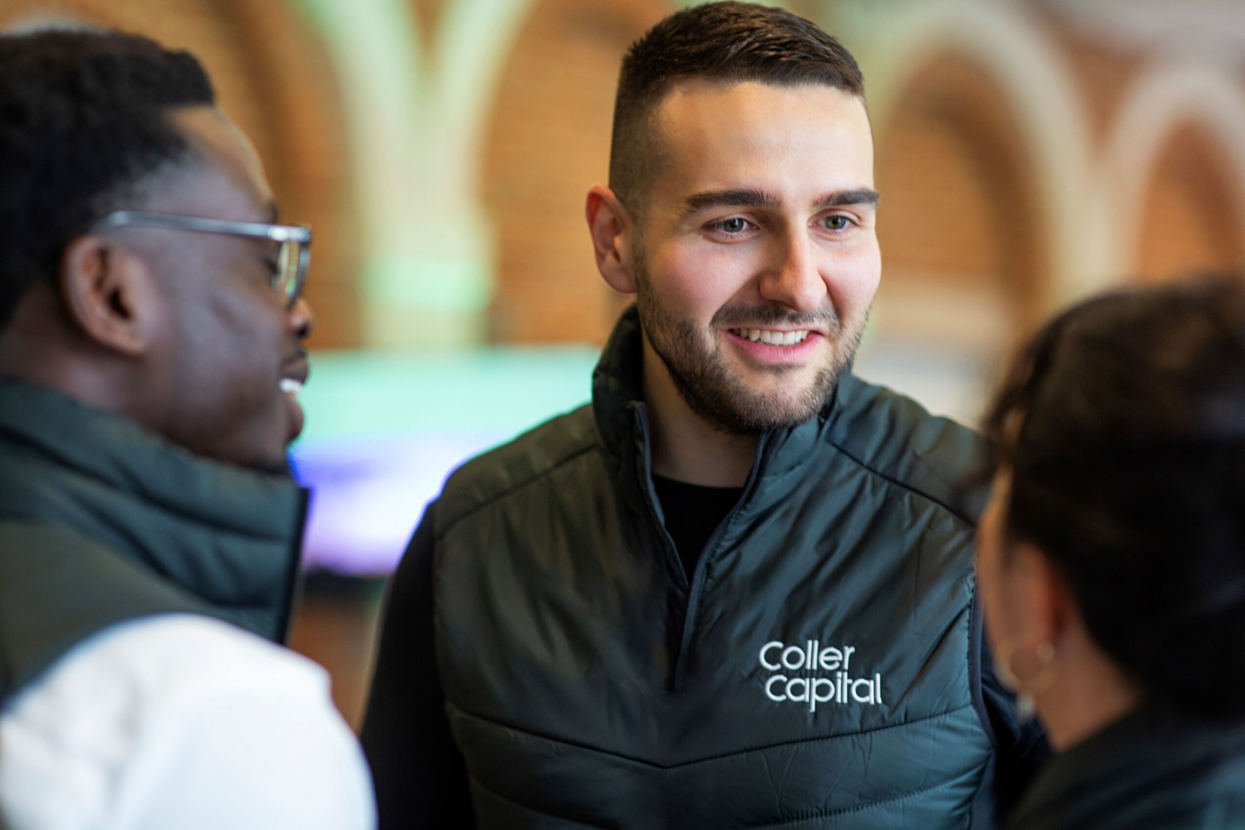 Three individuals are engaged in a conversation, wearing dark outerwear, with a warm brick background enhancing the atmosphere.
